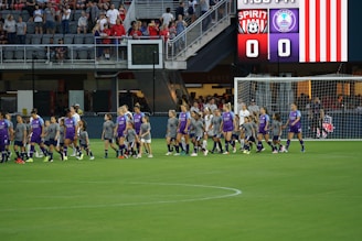 A group of soccer players in purple and gray uniforms are walking on a soccer field. Behind them, an electronic scoreboard displays the team names, Spirit and Orlando Pride, both with a score of 0. In the background, spectators are seated in the stands, watching the athletes.