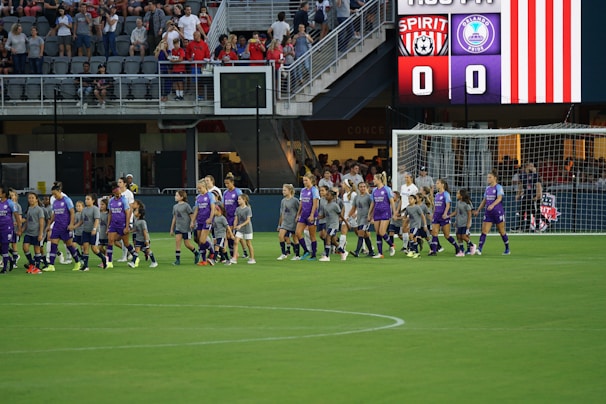 A group of soccer players in purple and gray uniforms are walking on a soccer field. Behind them, an electronic scoreboard displays the team names, Spirit and Orlando Pride, both with a score of 0. In the background, spectators are seated in the stands, watching the athletes.