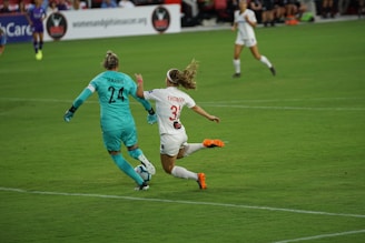Two soccer players are in action on the field, with one wearing a teal uniform and gloves, likely the goalkeeper, and the other in a white uniform with orange cleats. They are focused on the ball, which is not visible, indicating a dynamic moment in the match.