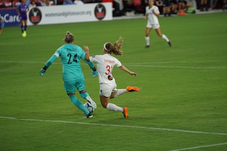Two soccer players are in action on the field, with one wearing a teal uniform and gloves, likely the goalkeeper, and the other in a white uniform with orange cleats. They are focused on the ball, which is not visible, indicating a dynamic moment in the match.