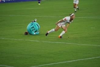 A soccer player in a white uniform with orange cleats is moving swiftly across the green grass field. Another player in a teal goalkeeper jersey with the number 24 is on the ground nearby, having fallen or dived. The scene suggests a moment of action during a soccer match.