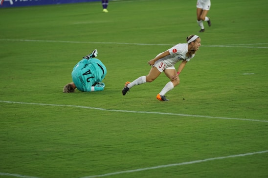 A soccer player in a white uniform with orange cleats is moving swiftly across the green grass field. Another player in a teal goalkeeper jersey with the number 24 is on the ground nearby, having fallen or dived. The scene suggests a moment of action during a soccer match.