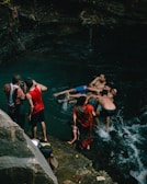 People are gathered at a natural pool or waterfall, with some in the water and others standing on rocks. The group includes men, women, and a child, and they're enjoying a leisurely time amidst nature.