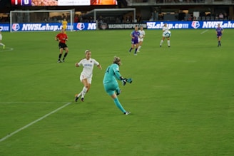 A women's soccer match is taking place on a well-maintained grass field. Players from two teams are actively engaged, with one team wearing white kits and the other in purple. A referee in red is also visible. A goalkeeper in a teal uniform is about to kick the ball as an opponent in white approaches.