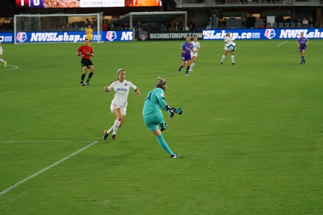 A women's soccer match is taking place on a well-maintained grass field. Players from two teams are actively engaged, with one team wearing white kits and the other in purple. A referee in red is also visible. A goalkeeper in a teal uniform is about to kick the ball as an opponent in white approaches.