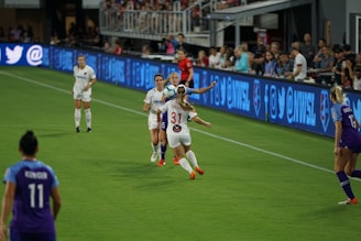 A women's soccer match is taking place with players from two teams in action. Players in white and purple uniforms are competing for the ball on a grassy field. Spectators are visible in the background watching the game, and advertisements are displayed on screens along the sidelines.
