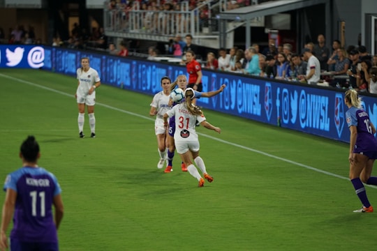 A women's soccer match is taking place with players from two teams in action. Players in white and purple uniforms are competing for the ball on a grassy field. Spectators are visible in the background watching the game, and advertisements are displayed on screens along the sidelines.