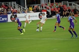 A soccer match in progress with players actively competing for the ball on a well-maintained field. The players are wearing team uniforms, and there is a crowd in the grandstand watching the game intently with some banners and advertisements visible in the background.