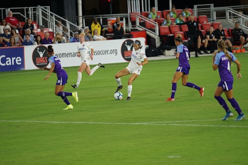 A soccer match in progress with players actively competing for the ball on a well-maintained field. The players are wearing team uniforms, and there is a crowd in the grandstand watching the game intently with some banners and advertisements visible in the background.