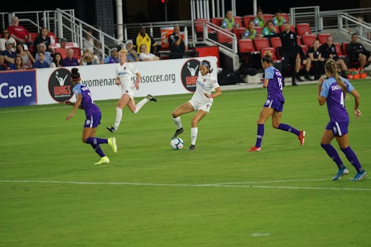 A soccer match in progress with players actively competing for the ball on a well-maintained field. The players are wearing team uniforms, and there is a crowd in the grandstand watching the game intently with some banners and advertisements visible in the background.