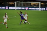 A soccer match with athletes actively engaged on the field. One player in a purple uniform is attempting to control the ball, while two players in white uniforms are nearby, one preparing to challenge for the ball. The background features a soccer goal and illuminated advertising boards.