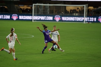 A soccer match with athletes actively engaged on the field. One player in a purple uniform is attempting to control the ball, while two players in white uniforms are nearby, one preparing to challenge for the ball. The background features a soccer goal and illuminated advertising boards.