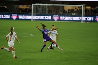 A soccer match with athletes actively engaged on the field. One player in a purple uniform is attempting to control the ball, while two players in white uniforms are nearby, one preparing to challenge for the ball. The background features a soccer goal and illuminated advertising boards.