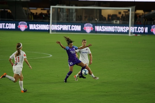 A soccer match with athletes actively engaged on the field. One player in a purple uniform is attempting to control the ball, while two players in white uniforms are nearby, one preparing to challenge for the ball. The background features a soccer goal and illuminated advertising boards.