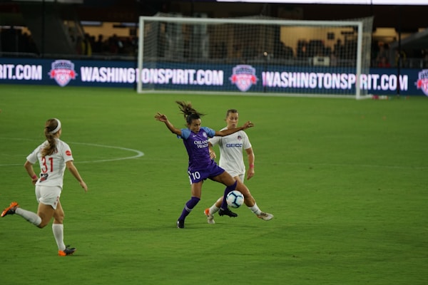 A soccer match with athletes actively engaged on the field. One player in a purple uniform is attempting to control the ball, while two players in white uniforms are nearby, one preparing to challenge for the ball. The background features a soccer goal and illuminated advertising boards.