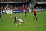 A soccer match is taking place on a well-maintained field. A player in a white jersey is falling forward, possibly after a physical interaction with a player in a purple jersey. Other players and a referee watch the event unfold. The stands are filled with spectators, and advertisements are visible along the sidelines.