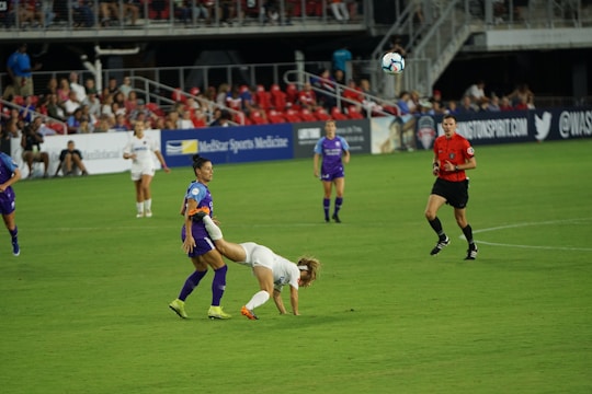 A soccer match is taking place on a well-maintained field. A player in a white jersey is falling forward, possibly after a physical interaction with a player in a purple jersey. Other players and a referee watch the event unfold. The stands are filled with spectators, and advertisements are visible along the sidelines.