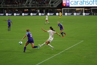 A soccer match is taking place on a grassy field. One player in a purple jersey is attempting to control the ball while an opposing player in a white jersey is trying to intercept. Other players are visible in the background, and there's an audience in the stands. The scoreboard shows a tied score, and advertisements are displayed around the field.