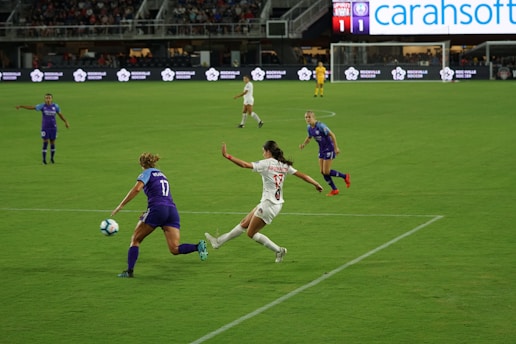 A soccer match is taking place on a grassy field. One player in a purple jersey is attempting to control the ball while an opposing player in a white jersey is trying to intercept. Other players are visible in the background, and there's an audience in the stands. The scoreboard shows a tied score, and advertisements are displayed around the field.