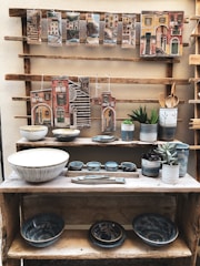 A rustic wooden shelf displaying various pieces of pottery including bowls, mugs, and decorative plaques. The plaques feature colorful, painted cityscapes with Italian place names. The pottery pieces are primarily in earthy tones with some blue accents. Several small green plants in pots are also placed on the shelves.