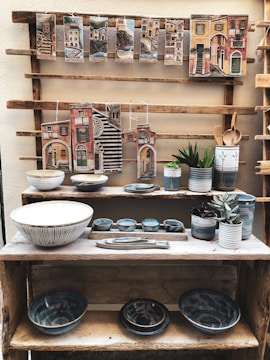 A rustic wooden shelf displaying various pieces of pottery including bowls, mugs, and decorative plaques. The plaques feature colorful, painted cityscapes with Italian place names. The pottery pieces are primarily in earthy tones with some blue accents. Several small green plants in pots are also placed on the shelves.