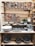 A rustic wooden shelf displaying various pieces of pottery including bowls, mugs, and decorative plaques. The plaques feature colorful, painted cityscapes with Italian place names. The pottery pieces are primarily in earthy tones with some blue accents. Several small green plants in pots are also placed on the shelves.