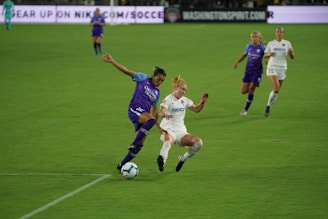 Two women engage in a competitive soccer match on a well-maintained grass field. One player, dressed in a purple uniform, attempts a maneuver against an opposing team member in a white uniform. The background features additional players and blurred advertisements on the stadium walls.