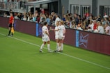 Two soccer players in white uniforms stand on the sideline of a field near a digital advertising board. One player holds a beverage bottle, and they appear to be in conversation. A referee in black stands nearby, and spectators fill the stands in the background, watching the event.