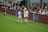 Two soccer players in white uniforms stand on the sideline of a field near a digital advertising board. One player holds a beverage bottle, and they appear to be in conversation. A referee in black stands nearby, and spectators fill the stands in the background, watching the event.