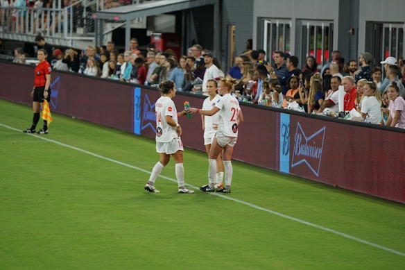 Two soccer players in white uniforms stand on the sideline of a field near a digital advertising board. One player holds a beverage bottle, and they appear to be in conversation. A referee in black stands nearby, and spectators fill the stands in the background, watching the event.