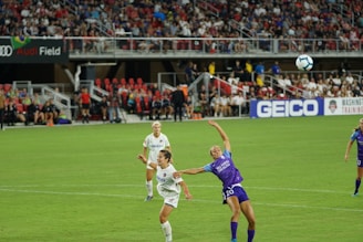 A women's soccer match is in progress on a well-maintained field with players actively competing for the ball. Two players, one in a white jersey and the other in a purple jersey, are the focal point as they leap towards the ball in mid-air. A lively crowd fills the stands in the background, with banners and advertisements visible, including 'Audi Field' and 'GEICO'.
