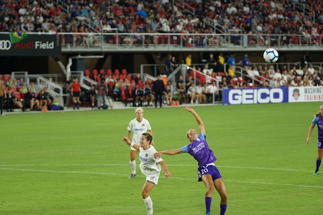 A women's soccer match is in progress on a well-maintained field with players actively competing for the ball. Two players, one in a white jersey and the other in a purple jersey, are the focal point as they leap towards the ball in mid-air. A lively crowd fills the stands in the background, with banners and advertisements visible, including 'Audi Field' and 'GEICO'.
