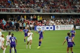 A women's soccer match is in action with teams wearing purple and white jerseys. Players are actively engaged with the ball mid-air, while a packed stadium full of spectators watches the event. The field is vibrant green, and a large advertisement banner is visible along the sidelines.