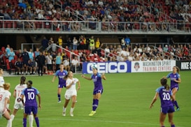 A women's soccer match is in action with teams wearing purple and white jerseys. Players are actively engaged with the ball mid-air, while a packed stadium full of spectators watches the event. The field is vibrant green, and a large advertisement banner is visible along the sidelines.