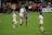 Two soccer players are competing for a header on the field during a match. One player, wearing a purple jersey, is jumping next to another player in a white jersey. Several other players and spectators are visible in the background, along with a vibrant green field.
