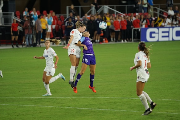 Two soccer players are competing for a header on the field during a match. One player, wearing a purple jersey, is jumping next to another player in a white jersey. Several other players and spectators are visible in the background, along with a vibrant green field.