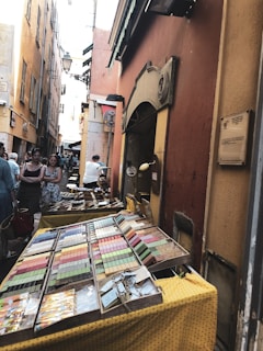 A narrow street lined with tall, closely packed buildings featuring a vibrant market stall selling colorful items placed on a yellow cloth-covered table. Several people are visible in the background, both browsing the stall and walking through the street. The overall setting gives off a European vibe, possibly in a historic city area.
