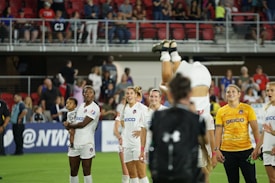 A group of female soccer players, some in white and one in a yellow jersey, are on a sports field. One player is mid-flip, causing excitement and anticipation among the team members. A player is holding a toddler in her arms, and there are spectators in the background in a stadium setting.
