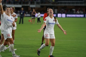 Several soccer players are on a field, celebrating with smiles and raised arms. They are wearing white jerseys with sponsor logos. The background shows a stadium with other people and advertisements.
