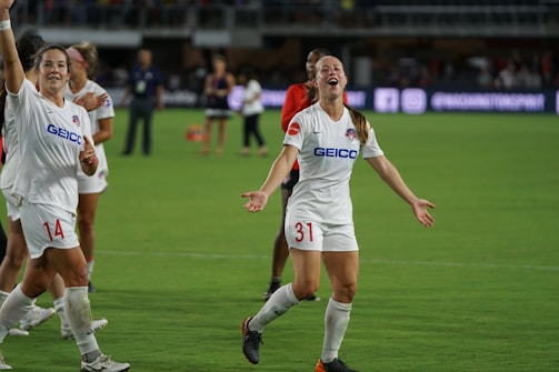 Several soccer players are on a field, celebrating with smiles and raised arms. They are wearing white jerseys with sponsor logos. The background shows a stadium with other people and advertisements.