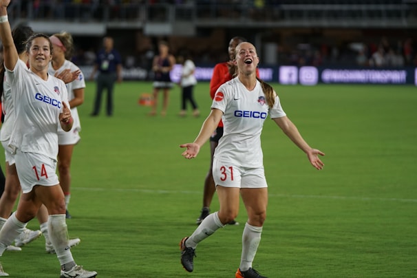 Several soccer players are on a field, celebrating with smiles and raised arms. They are wearing white jerseys with sponsor logos. The background shows a stadium with other people and advertisements.
