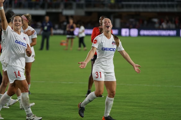 Several soccer players are on a field, celebrating with smiles and raised arms. They are wearing white jerseys with sponsor logos. The background shows a stadium with other people and advertisements.