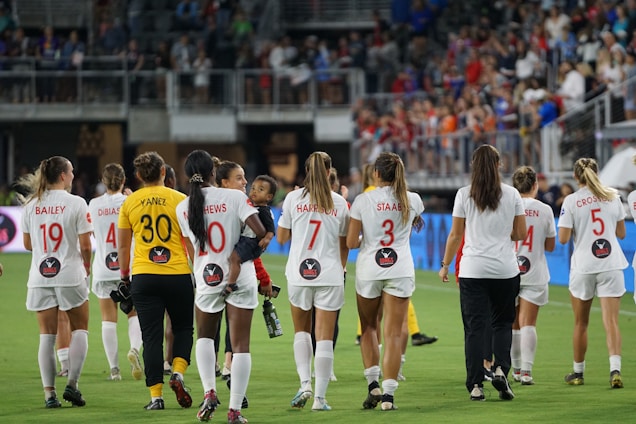 A group of female soccer players in matching uniforms, with numbers and names on their backs, walk together on a soccer field. They appear to be leaving the field, and some are holding or interacting with a child. The background shows a stadium with a crowd of spectators.