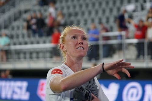 A person with light red hair is wearing a white sports jersey and has a towel draped over one arm. They appear to be gesturing or waving, with a serious or focused expression on their face. The background features an out-of-focus audience and stadium seating, suggesting a sports event.