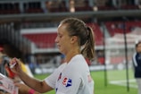 A female athlete with a ponytail and wearing a white sports jersey is signing an item, likely a piece of merchandise or fan memorabilia, with a marker. There are blurred figures and a partially visible sports field or stadium background, suggesting a post-game or practice setting.