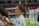 A female athlete with a ponytail and wearing a white sports jersey is signing an item, likely a piece of merchandise or fan memorabilia, with a marker. There are blurred figures and a partially visible sports field or stadium background, suggesting a post-game or practice setting.