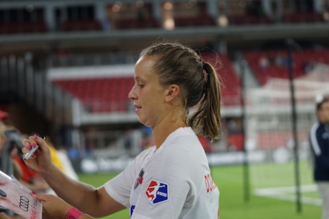 A female athlete with a ponytail and wearing a white sports jersey is signing an item, likely a piece of merchandise or fan memorabilia, with a marker. There are blurred figures and a partially visible sports field or stadium background, suggesting a post-game or practice setting.