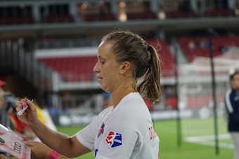 A female athlete with a ponytail and wearing a white sports jersey is signing an item, likely a piece of merchandise or fan memorabilia, with a marker. There are blurred figures and a partially visible sports field or stadium background, suggesting a post-game or practice setting.