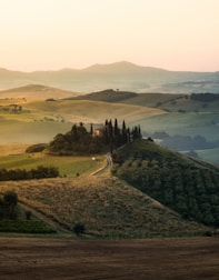 Rolling hills stretch into the distance under a soft, golden sunset glow. A winding path leads to a cluster of tall cypress trees surrounding a secluded villa. The landscape is dotted with fields of varying shades of green and brown, creating a patchwork effect. Distant mountains are visible on the horizon, adding depth to the serene countryside scene.