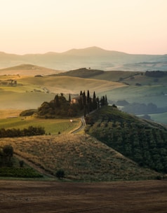 Rolling hills stretch into the distance under a soft, golden sunset glow. A winding path leads to a cluster of tall cypress trees surrounding a secluded villa. The landscape is dotted with fields of varying shades of green and brown, creating a patchwork effect. Distant mountains are visible on the horizon, adding depth to the serene countryside scene.